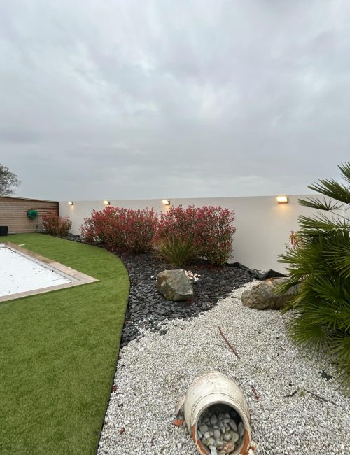 Installation de 4 luminaires extérieurs dans un jardin moderne à Saint-Julien-de-Concelles (44450), réalisée par Pierre Martienne, électricien. Les éclairages sont fixés sur un muret blanc et mettent en valeur les massifs paysagers et les plantes bordant une piscine.
